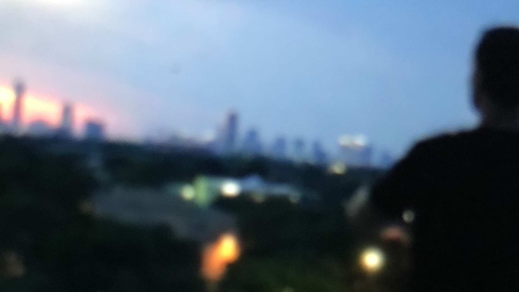 Blurry view of a city skyline at dusk with a silhouette of a person in the foreground, vibrant colors from the sunset and urban lights in the distance